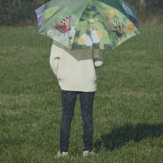Une personne portant un pull en tricot et un pantalon noir se tient sous un parapluie vert à motif de papillon sous la pluie.