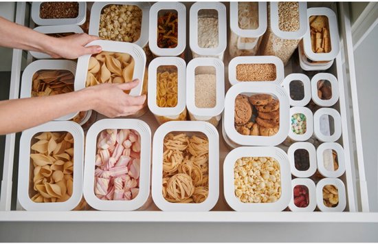 Un tiroir de cuisine rempli de boîtes de conservation Modula Mepal avec pâtes, pop-corn, biscuits et bonbons. Des mains attrapent une boîte de coquillettes.