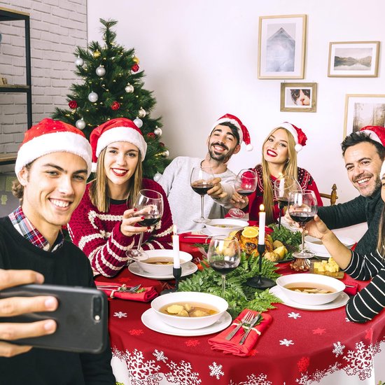 Gezellig kerstfeest met mensen die een selfie maken. De tafel is gedekt met een rood kersttafelkleed met witte sneeuwvlokken en bloemenprint.