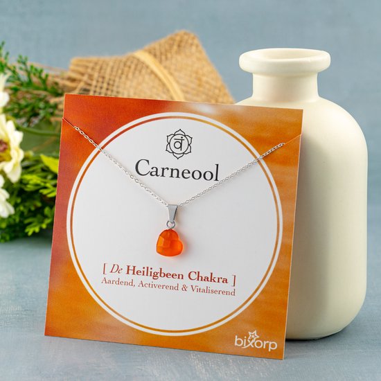 Zilveren ketting met hartvormige, oranje carneool hanger op een kaart met de tekst 'Carneool - [De Heiligbeen Chakra]'.