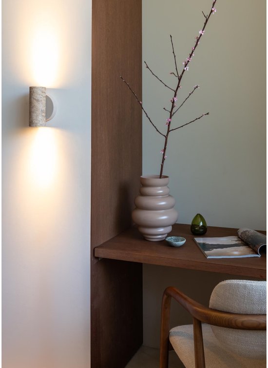 Een beige wandlamp van steen met twee lichtbundels op een bureau met vaas, tak, flesje en tijdschrift.