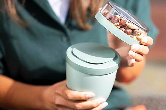 Vrouw vult een lichtgroene yoghurtbeker met noten uit een transparante deksel.