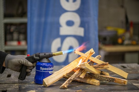 Een hand met een grijze handschoen houdt een blauwe gasbrander met vlam vast, die houten takjes aansteekt.
