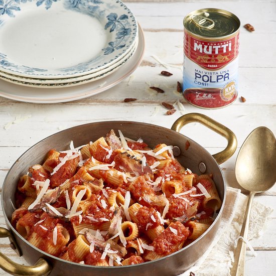 Koperen pan met rigatoni pasta in tomatensaus, gegarneerd met ansjovis, spek en Parmezaanse kaas. Naast een blik Mutti Polpa.