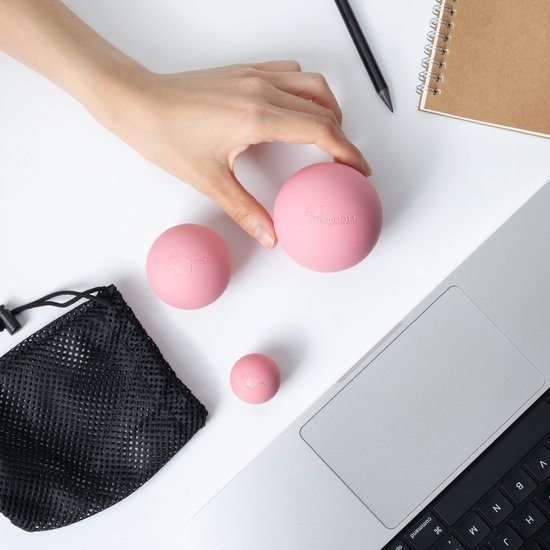 Een hand pakt een roze massagebal met logo, met twee kleinere roze ballen en een zwarte mesh tas ernaast, op een witte achtergrond.