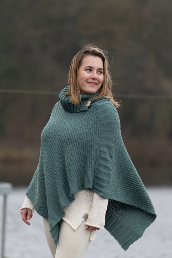 Groenblauwe gebreide poncho met hoge kraag en off-white onderlaag, gedragen door een vrouw met blond haar bij water.