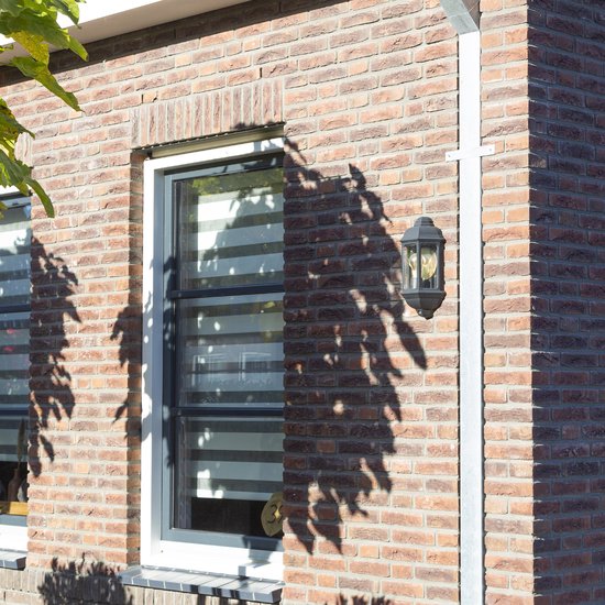 Een groene klassieke buitenlamp met een glazen kap hangt naast een raam met horizontale lamellen.