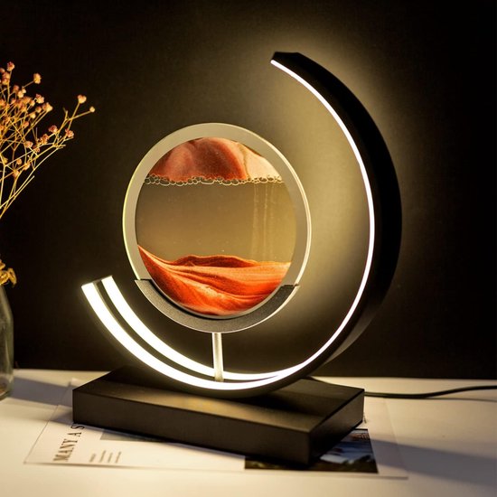 Rode zandkunst in een rond glas met een zwarte lamp, op een bureau naast een vaas met bloemen.