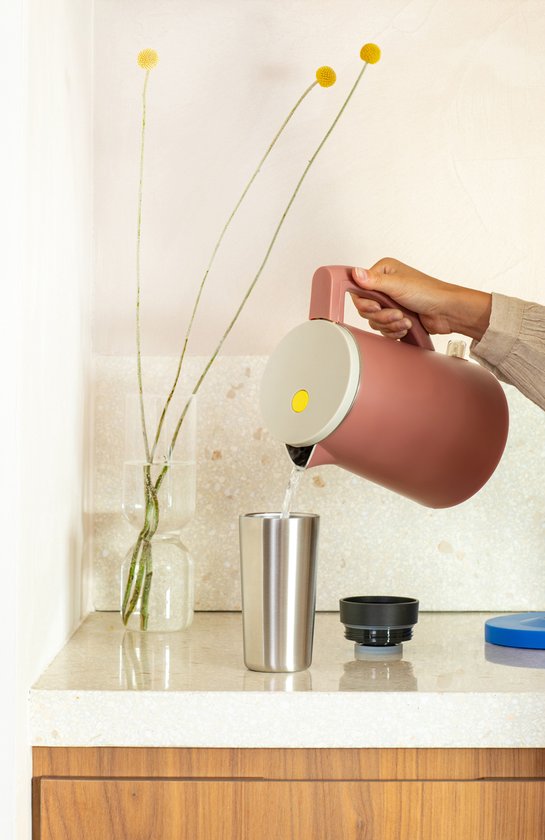Hand schenkt water uit een roze waterkoker in een roestvrijstalen beker met een zwarte deksel ernaast.