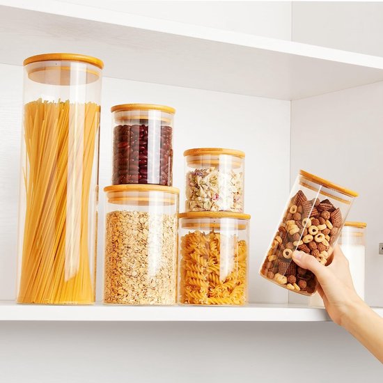 Glazen voorraadpotten met bamboedeksels, gevuld met spaghetti, ontbijtgranen, pasta en snacks, op een witte schap.