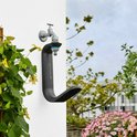 Zwarte Gardena Liano TapFix slanghouder aan een witte muur met kraan en tuinslang. Groene plant links, bloemen en terras rechts.