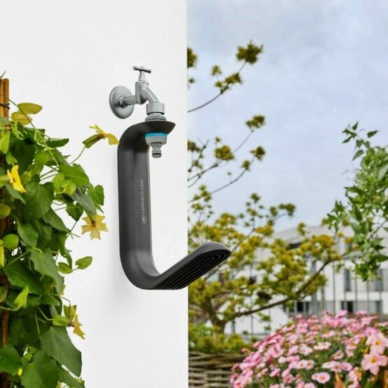 Zwarte Gardena Liano TapFix slanghouder aan een witte muur met kraan en tuinslang. Groene plant links, bloemen en terras rechts.