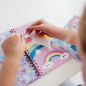 Une fille colle un autocollant étoile scintillant sur une licorne dans un livre "Sticker by number" Licorne. La page montre une licorne sur un arc-en-ciel.