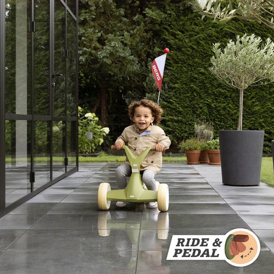 Vrolijk kind in beige jasje rijdt op een groene loopauto met beige zitting en handvatten, op een betegeld terras.