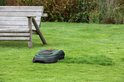 Groene en zwarte robotmaaier op een gazon naast een houten bankje.
