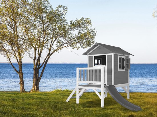 Grijs houten speelhuis op palen met witte accenten, veranda, trap en glijbaan, gefotografeerd met bomen en meer op de achtergrond.