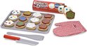 Houten koekjes met witte, chocolade, blauwe en groene icing, sprinkles en stippen op bakplaat met rode ovenwanten en spatel.