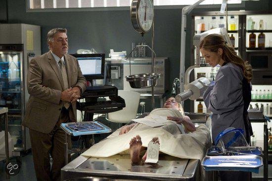 Dr. Maura Isles in grijze jas onderzoekt een lichaam op een tafel. Collega kijkt toe in mortuarium.