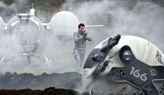 Tom Cruise in grijs pak met wapen, een beschadigde helm op de voorgrond.