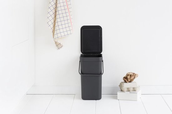 Poubelle Brabantia gris foncé avec couvercle entrouvert, dans une cuisine blanche.