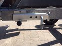 Zilverkleurige kantelbare steunpoot met ketting, bevestigd aan de zijkant van een aanhangwagen.
