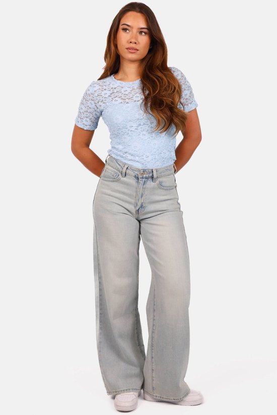 Model ten voeten uit in een lichtblauwe kanten top met korte mouwen, wijde spijkerbroek en witte sneakers.