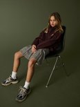 Model zittend in een bordeauxrode hoodie, geruite rok, witte sokken en sneakers tegen een groene achtergrond.