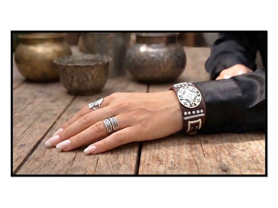 Bruin lederen armband met zilverkleurige studs en een rond, geborduurd detail. Gedragen om een vrouwenpols, samen met meerdere ringen.