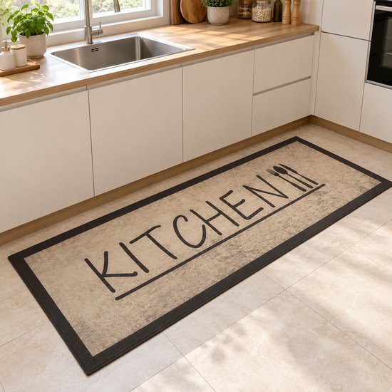 Beige keukenloper met zwarte rand en 'KITCHEN' opdruk ligt voor een keukenkastje op een tegelvloer.
