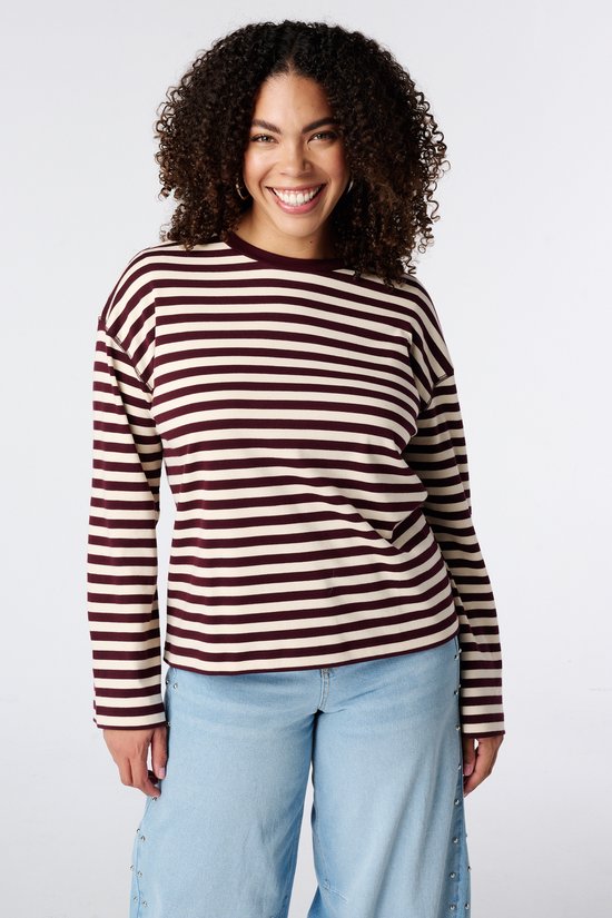 Model met krullend haar draagt een bordeaux en beige gestreepte longsleeve met ronde hals en lichtblauwe wijde jeans.