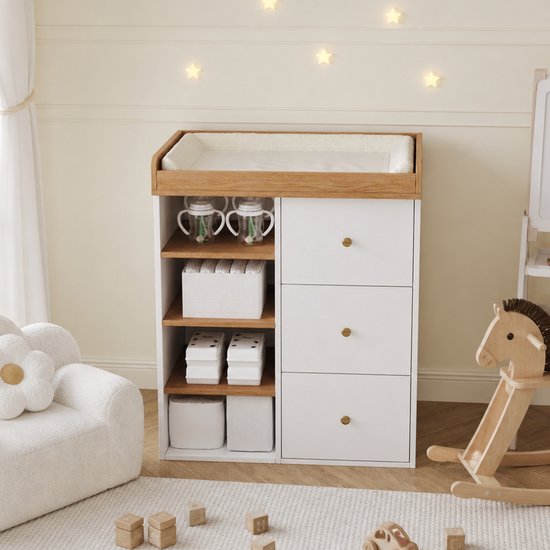 Wit-houten babycommode met drie lades en open legplanken, geplaatst in een kinderkamer met houten schommelpaard.