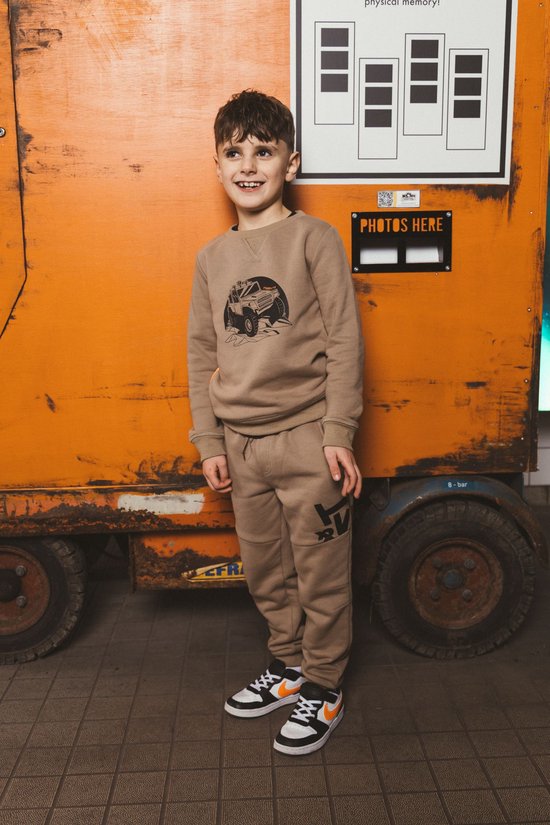 Jongen draagt een beige sweater en joggingbroek met Tygo & Vito print, staand voor een oranje achtergrond.