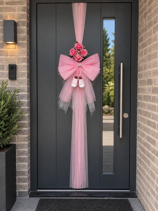 Roze geboortestrik met tule, kunstrozen en babyschoentjes aan een voordeur.