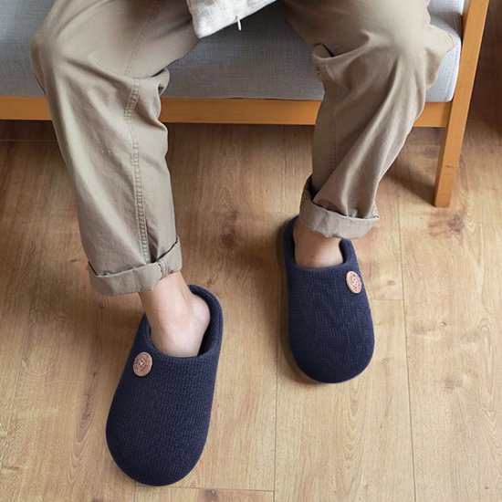 Persoon draagt zwarte, geribbelde pantoffels met decoratieve knoop terwijl ze op een stoel zit.