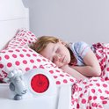 Een slapend meisje in bed, naast een wit schaapwekker met rode wijzerplaat. Ze ligt onder een dekbed met roze stippen.