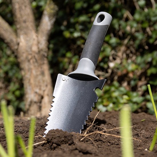 Gekartelde zwarte spade met grijze handgreep steekt uit de aarde, met achtergrond van boom en groen.