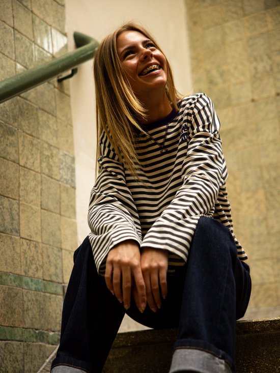 Een lachende jonge vrouw met bretels, zittend op een trap, met een gestreept longsleeve shirt aan.