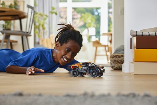 Kind speelt met een oranje en zwarte LEGO Technic NEOM McLaren Extreme E Team auto op een houten vloer.