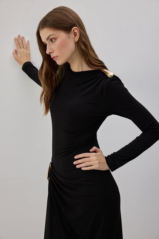 Zijaanzicht van een model in een zwarte maxi-jurk met gouden schouder- en heupdetails, leunend tegen een muur.