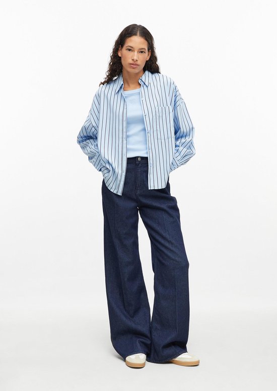 Model in lichtblauw geribbeld shirt onder een gestreept overhemd met donkere jeans.