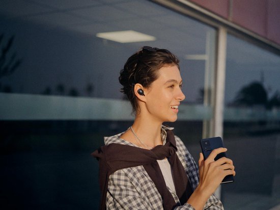 Jonge vrouw met oordopjes in, houdt smartphone vast, glimlacht naar rechts.