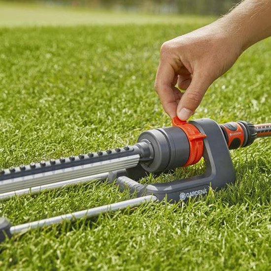 Een hand stelt de oranje hendel in op de Gardena zwenksproeier, liggend op groen gras.