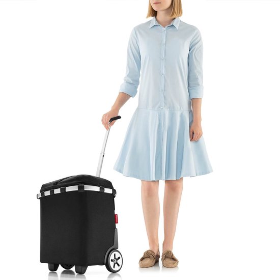 Een vrouw met een lichtblauwe jurk staat naast een
zwarte Reisenthel boodschappentrolley met wielen en
uitgeschoven handgreep.