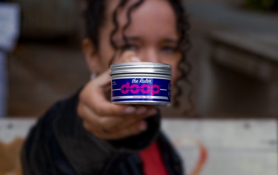 Een persoon met krullend haar houdt een zilverkleurige pot met de tekst 'the Ruler doop SHAPING PASTE' voor de camera.