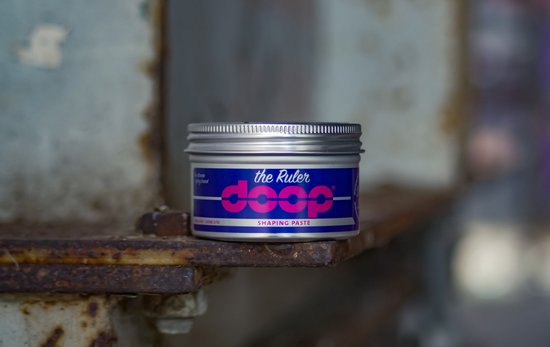 Zilverkleurige pot met de tekst 'the Ruler doop SHAPING PASTE' op een blauw label met roze letters.