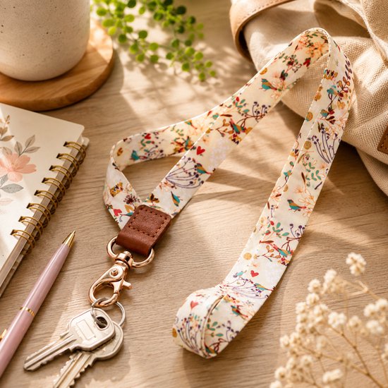 Crèmekleurige lanyards met vogel- en hartjesprint, vastgemaakt aan sleutels op een houten bureau, naast een notitieboekje.