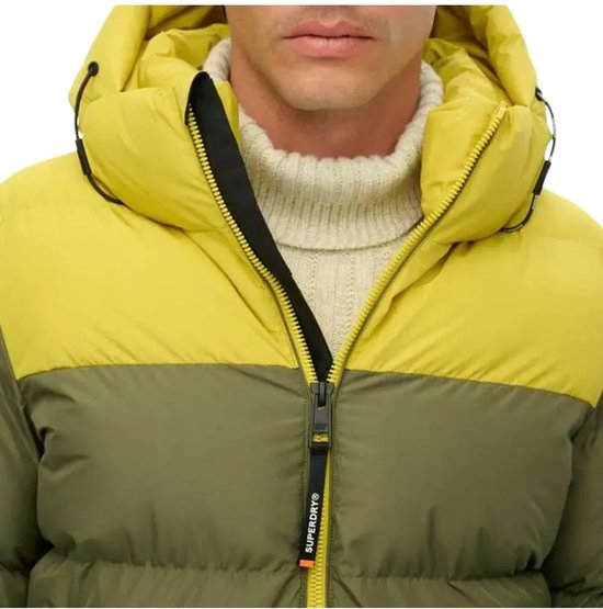 Close-up van een tweekleurige Superdry jas in olijfgroen en geel met 'Superdry' op de ritssluiting.