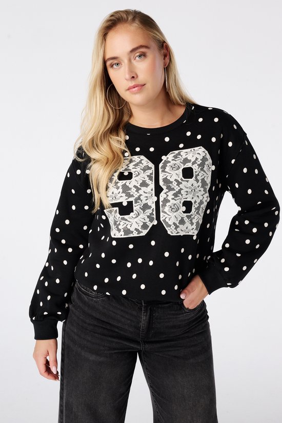 Model draagt een zwarte sweater met witte stippen en '98' in wit kant, hand in de zak.