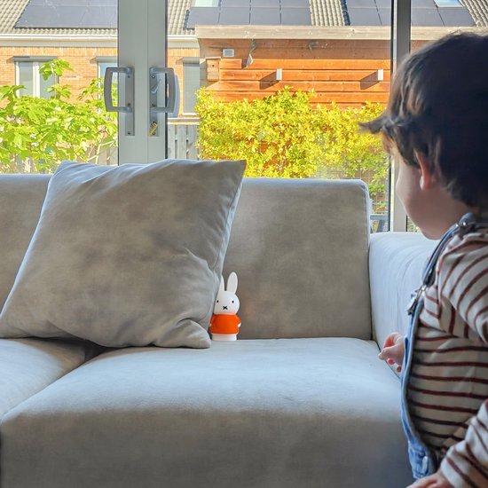 Kind kijkt naar een Nijntje-lampje in oranje outfit dat verstopt staat achter een kussen op de bank.