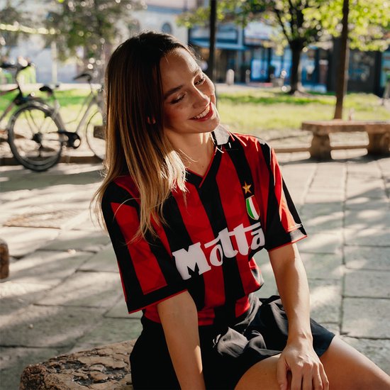Een glimlachende vrouw draagt een retro rood-zwart voetbalshirt met het 'Motta'-logo in een park.